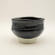 A handmade ceramic tea bowl with a glossy black glaze and a contrasting light beige unglazed base, photographed against a neutral off-white background.