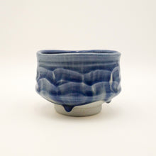 A handmade ceramic tea bowl with a deep blue glaze and textured, wavy relief patterns, featuring a small protruding tab on the front and a light gray unglazed base, set against a plain off-white background.