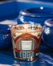 A lit Baobab Collection 'Mamakech' scented candle in a clear glass jar with a colorful Moroccan arch pattern, set against a background of blue ceramic vessels.