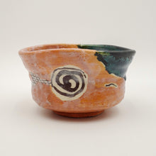 A handmade ceramic bowl with a terracotta orange body, a dark green glazed interior, and a black and white spiral motif on the side, set against a plain light gray background.