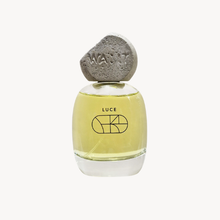 A clear glass perfume bottle filled with pale yellow liquid, featuring a textured gray stone-like cap embossed with the word 'WA:IT'. The bottle is labeled 'LUCE' with a minimalist geometric line graphic.