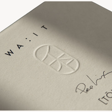 Close-up of a textured off-white paper surface with embossed branding, including the word 'WAIT' in black letters, a white oval monogram logo, and a cursive signature.