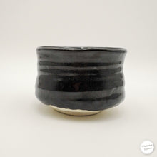 A handmade black glazed ceramic tea whisking bowl with horizontal ridges and a natural beige foot ring, photographed on a clean white background.