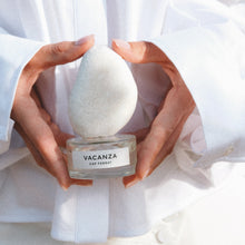A person in a white shirt holds a perfume bottle labeled 'VACANZA CAP FERRAT' with a white teardrop-shaped cap on a bright, sunlit background.