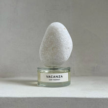 A perfume bottle with a white label reading 'VACANZA CAP FERRAT' sits on a light gray surface against a matching gray background, with a smooth white stone sculpture balanced on top.