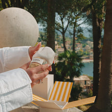A person in a white jacket holds a perfume bottle labeled 'VACANZA' in front of an open striped box, with a scenic outdoor background of trees, water, and buildings.