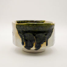 A handmade ceramic tea bowl with a textured off-white body, featuring a glossy dark green and black glaze drip design on the front, set against a plain light gray background.