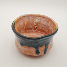 A handmade ceramic bowl with a terracotta orange body, featuring a dark teal-green glazed rim with dripping paint and a small green accent on the side, set against a plain light background.