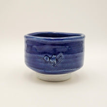 A small handmade ceramic tea bowl with a glossy cobalt blue glaze and a textured, hand-painted motif on the front, resting on a light gray base against a neutral off-white background.