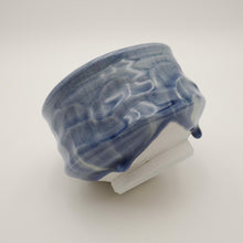 A handmade ceramic bowl with a glossy blue and white marbled glaze, resting on a white rectangular display block against a neutral light gray background.