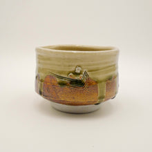 A handmade ceramic tea bowl with a rustic, hand-painted design featuring a small white bird and a brown landscape, covered in a glossy olive-green and amber-brown glaze, set against a plain white background.