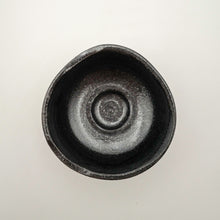 A top-down view of a handmade ceramic bowl with a dark, textured glaze on a plain off-white background.