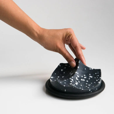 Hand interacting with a black ceramic dish on a light gray background