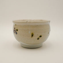 A handmade ceramic tea bowl with a speckled beige glaze and dark brown and green spot accents, photographed against a neutral light gray background.