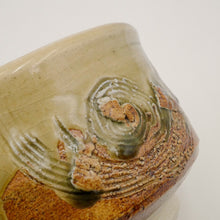 Close-up of a handmade ceramic bowl with a textured, organic spiral design in earthy brown and olive green glazes over a cream base, on a plain white background.