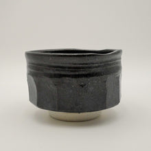 A handmade ceramic tea bowl with a dark charcoal black glaze and a contrasting light beige unglazed foot ring, photographed against a neutral light gray background.
