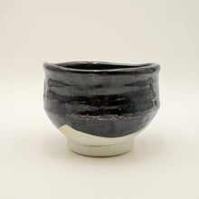 A handmade ceramic tea bowl with a glossy black glaze on the upper half and a matte off-white base on the lower half, photographed against a plain light background.