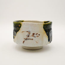 A handmade ceramic tea bowl with a matte off-white glaze, dark green and black dripped glaze accents, and brown hand-painted calligraphic characters on the front, set against a plain white background.