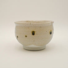 A handmade ceramic tea bowl with a speckled off-white glaze and dark brown spots, photographed against a neutral light gray background.