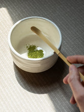 Person using a wooden spoon to mix green matcha powder in a white bowl on a textured surface.