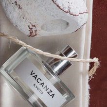 A clear glass perfume bottle labeled 'VACANZA KYOTO' with a silver atomizer cap, wrapped in natural twine, resting on a beige fabric surface next to a textured stone.