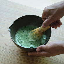 A person's hands whisking green matcha tea in a dark ceramic bowl using a bamboo whisk on a light wood table.