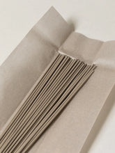 A close-up of thin, light brown incense sticks arranged inside a folded sheet of kraft paper on a plain off-white background.