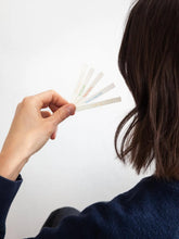 A person holding a fanned-out set of white paper incense strips with small colored text, shown from behind against a plain light background.