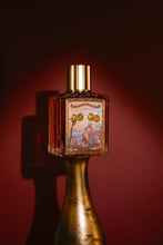 A decorative perfume bottle with a gold cap and a colorful illustrated label, mounted on a metallic gold base against a dark red background.