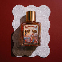 A rectangular glass perfume bottle with a gold cap and an ornate illustrated label, placed on a white scalloped paper doily against a dark red background.