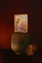 LT Piver NOHIBA box standing upright in a dark textured ceramic vase, with a gold-toned decorative flower prop on the surface beside it. The box features an illustrated scene with gold circular emblems and a multicolored border.