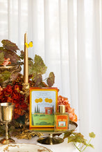 Decorative table setting with a candle, flowers, and a bottle of perfume on a white curtain background.