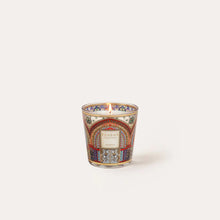 A small decorative candle in a patterned glass vessel with a white label, photographed against a plain light beige background.