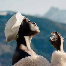 A person outdoors wearing a cream beret and cream fuzzy jacket, holding a small perfume bottle near their face with mountains blurred in the background.