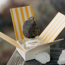 A perfume bottle labeled 'VACANZA MEXICO' sits inside an open white box with yellow-and-white striped flaps. A dark porous rock is placed on top of the bottle, and a few clear ice crystals rest on the wooden surface nearby.