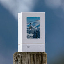 A white rectangular product box with a printed scenic mountain-and-ski-lift image on the front, displayed upright on a wooden post against a blue sky background.
