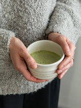 A person in a cozy gray knit sweater holds a small white ceramic cup filled with green matcha tea.