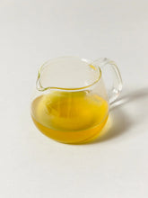 A small clear glass pitcher filled with golden yellow tea, photographed on a plain light background.