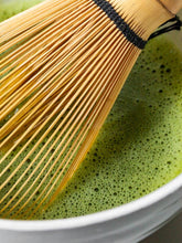 Wooden whisk being used to stir green tea in a white bowl