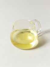 A small clear glass pitcher filled with pale yellow liquid, photographed on a clean light gray background.