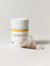 A white cylindrical canister of MORIHATA tea with a single triangular  teabag placed in front on a light gray studio background.