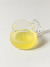 A clear glass pitcher filled with pale yellow tea, photographed on a clean white background with a soft shadow.