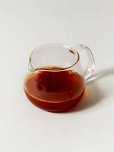 A small clear glass pitcher filled with dark amber-brown tea, photographed on a clean light gray background.