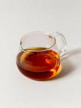A clear glass pitcher filled with amber-brown tea, photographed on a clean light gray background.