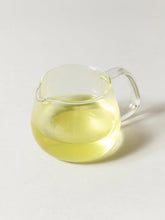 A clear glass pouring pitcher filled with pale yellow liquid, photographed on a clean light gray background.