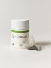 A white cylindrical container of MORIHATA tea with a green stripe and logo, shown next to a single triangular tea bag on a light gray background.