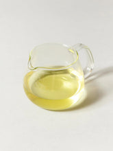 A clear glass pitcher filled with pale yellow liquid, photographed on a clean light gray background.