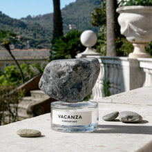 A perfume bottle labeled 'VACANZA PORTOFINO' with a large dark gray stone cap balancing on its rim, set on a stone terrace outdoors with hills and classical architecture in the background.