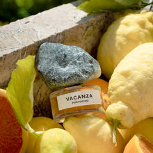 A perfume bottle labeled 'VACANZA PORTOFINO' sits on a rough gray stone, surrounded by lemons, a sliced orange, and a stone edge in an outdoor setting.