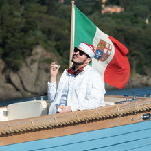 A man wearing a white jacket, sunglasses, and a red-and-white hat sits in a wooden boat on the water, with a large Italian flag flying behind him and a rocky, tree-covered shoreline in the background.